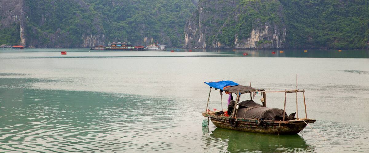 A traditional boat with a blue tarp on calm water surrounded by lush green mountains.