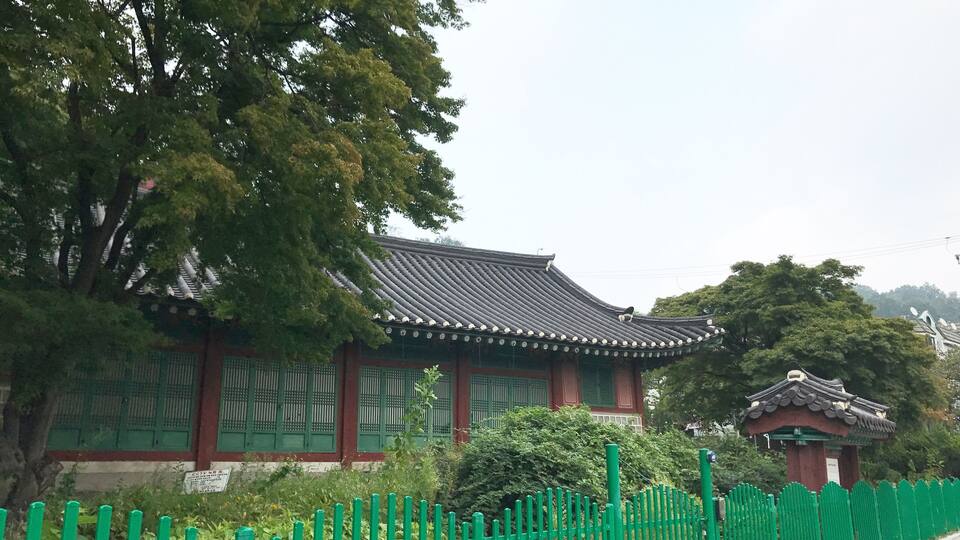A traditional Korean building with a tiled roof and green wooden windows, surrounded by lush trees and a green fence.