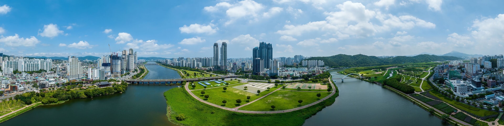 Um rio serpenteia por uma cidade com edifícios altos e modernos, uma ponte e áreas verdes.