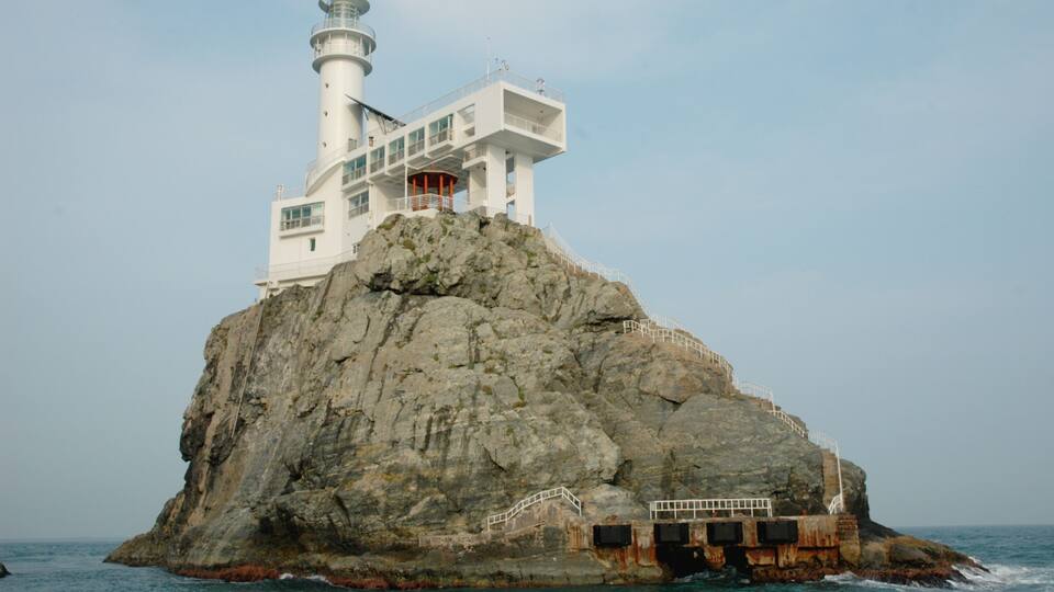 A lighthouse on a rocky outcrop with a white building on top, surrounded by the sea.