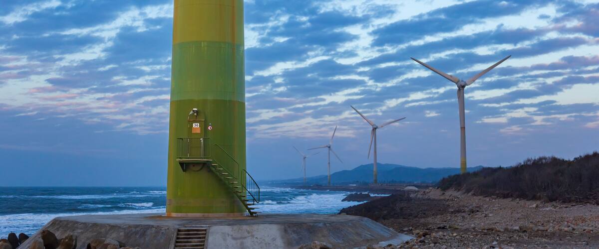 A green and yellow lighthouse by the sea with wind turbines in the background.