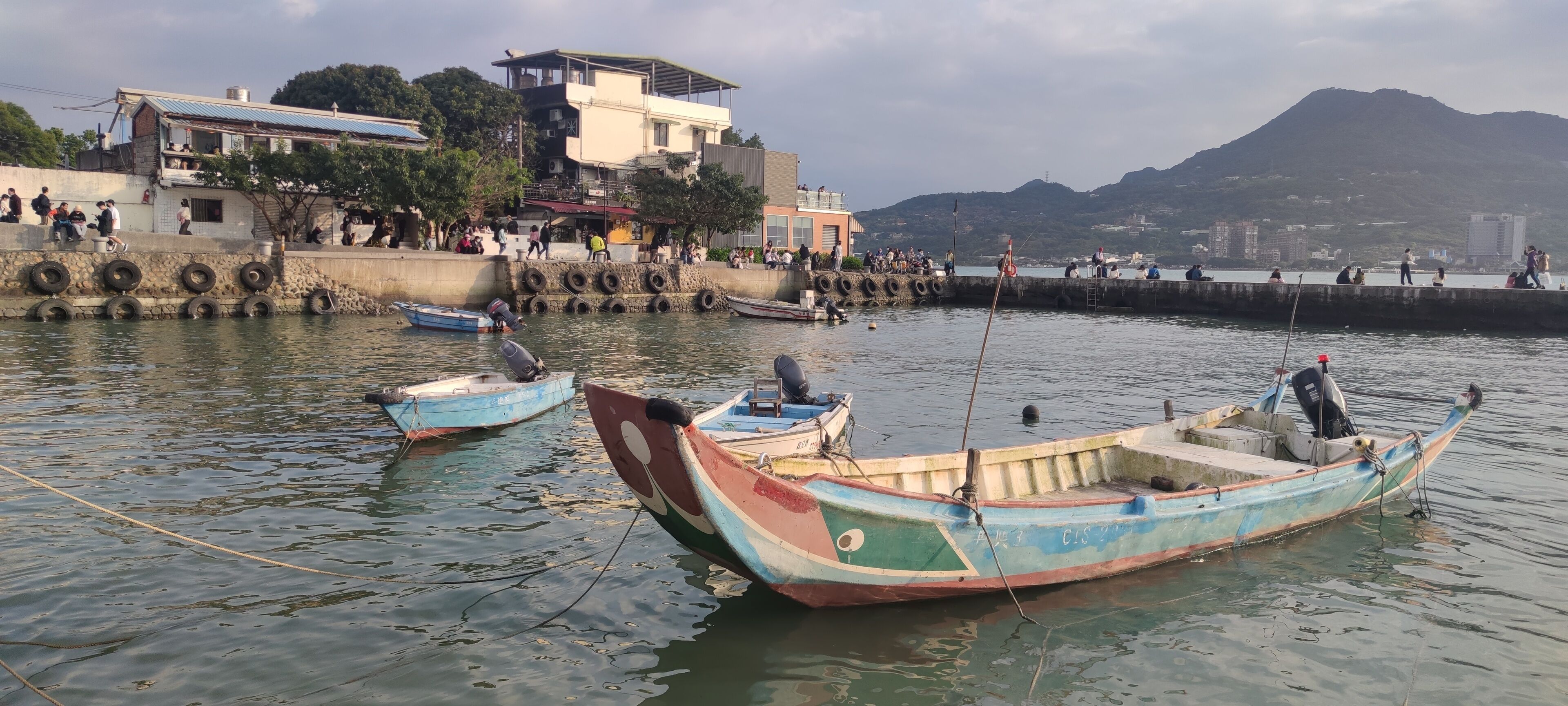 A harbor with boats, a stone wall with tires, and buildings in the background.