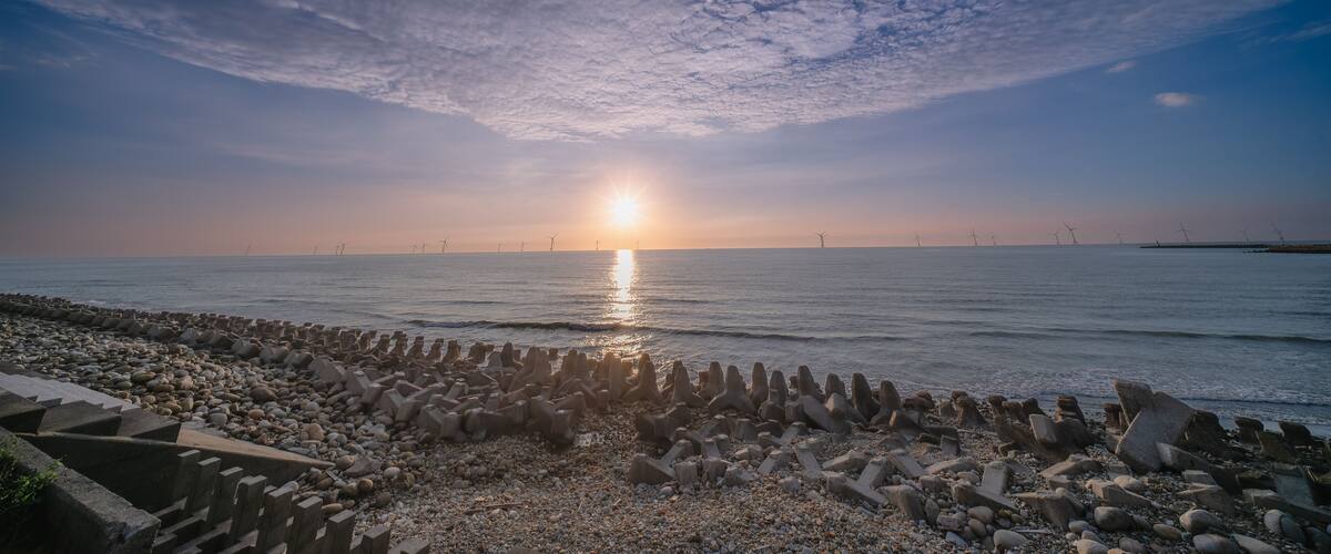 A sunset over a rocky shoreline with wind turbines in the distance.