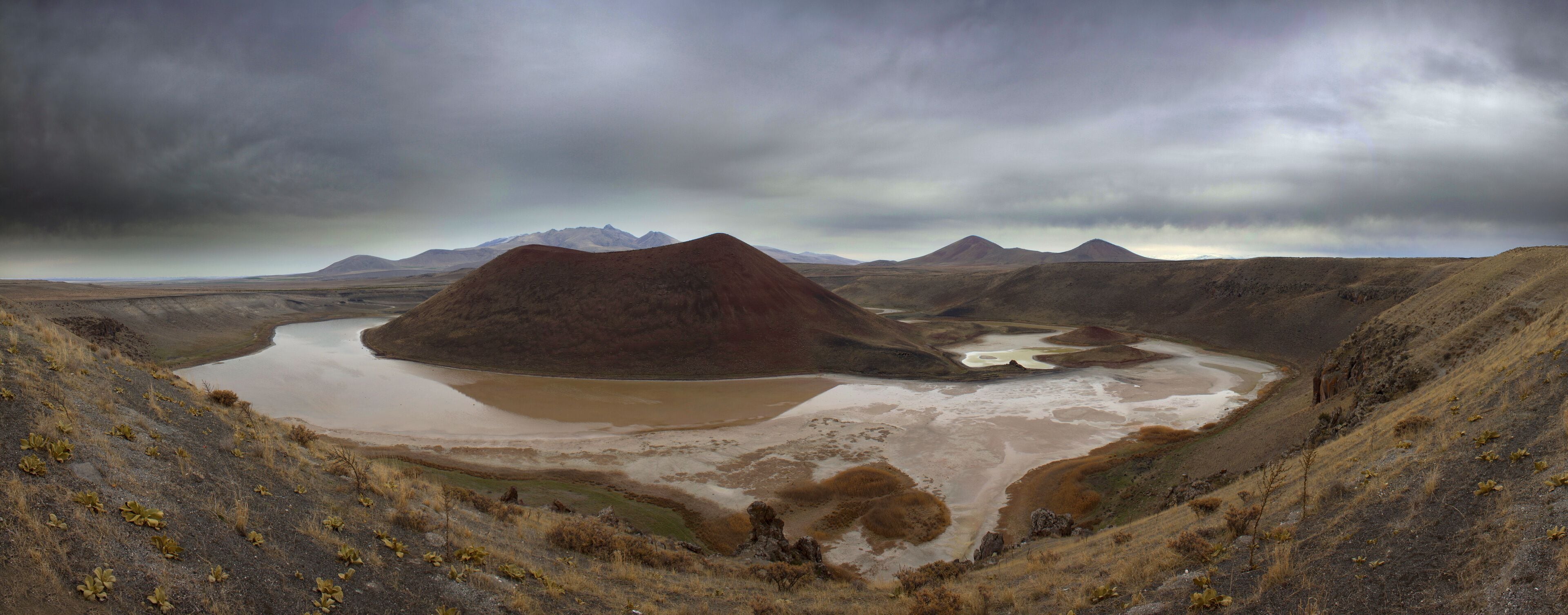 A landscape with a large lake surrounded by hills and mountains under a cloudy sky.