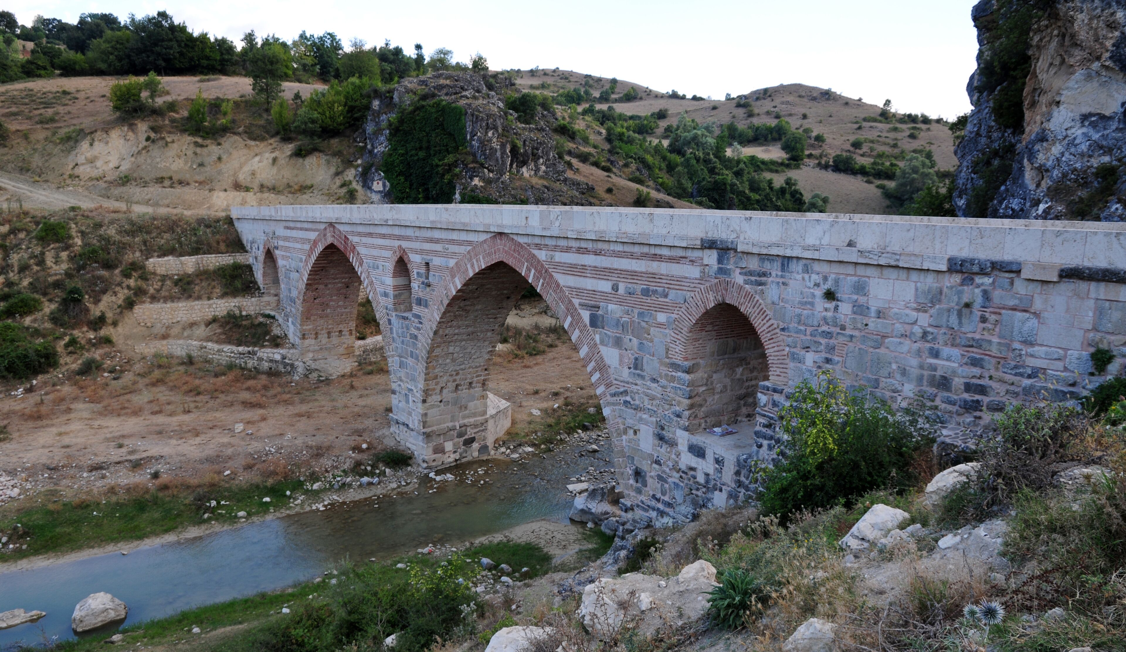 A stone bridge with three arches over a river, surrounded by hills and sparse vegetation.