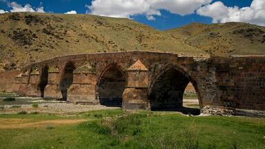 A stone bridge with multiple arches over a river, set against a backdrop of hills and a clear blue sky.