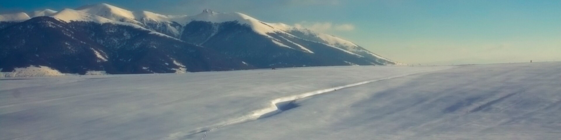 Paisagem coberta de neve com uma cordilheira ao fundo e céu claro.