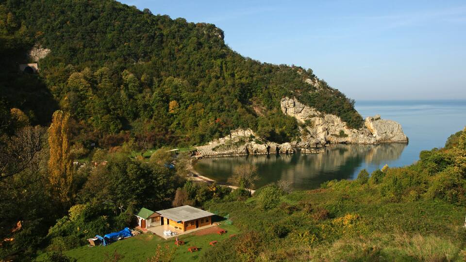 A coastal landscape with a house on a grassy hill, a rocky cliff, and a calm sea.
