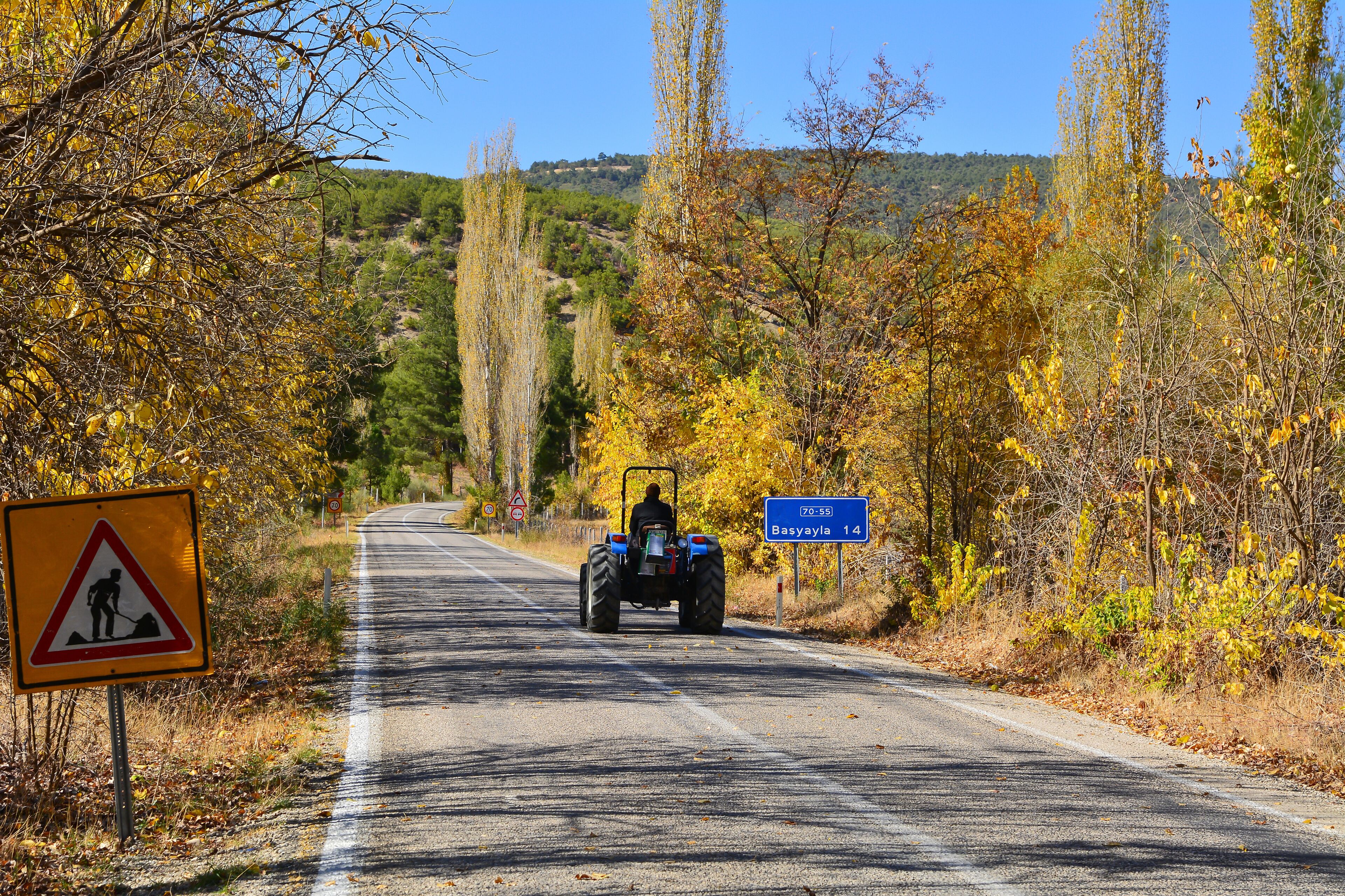 A rural road with a tractor, autumn foliage, and a construction warning sign.