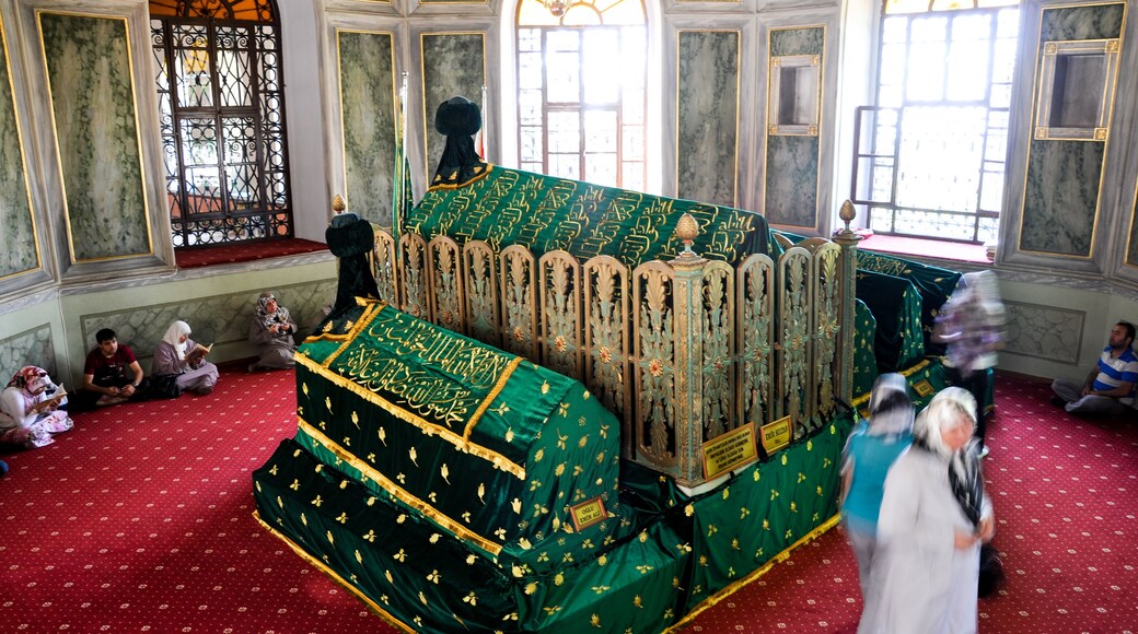 Una habitación con muebles ornamentados, un tapiz verde y dorado cubriendo una pieza central, y personas sentadas en el suelo.