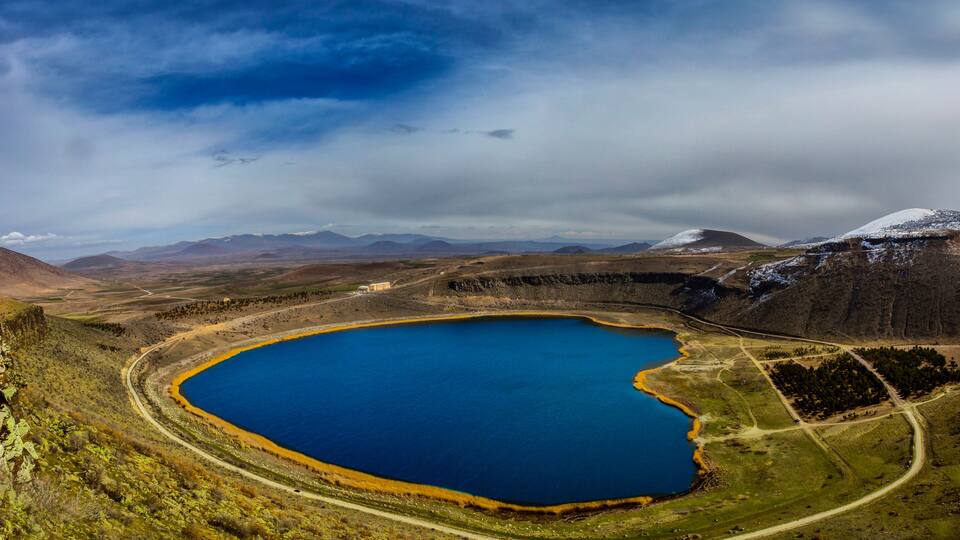 A large, circular lake surrounded by mountains and a cloudy sky.