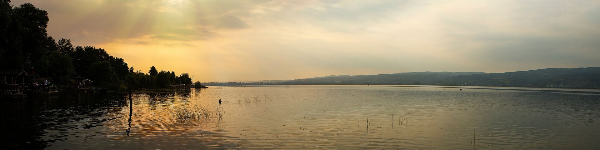 Un lago tranquilo con reflejos de árboles y un puente distante.