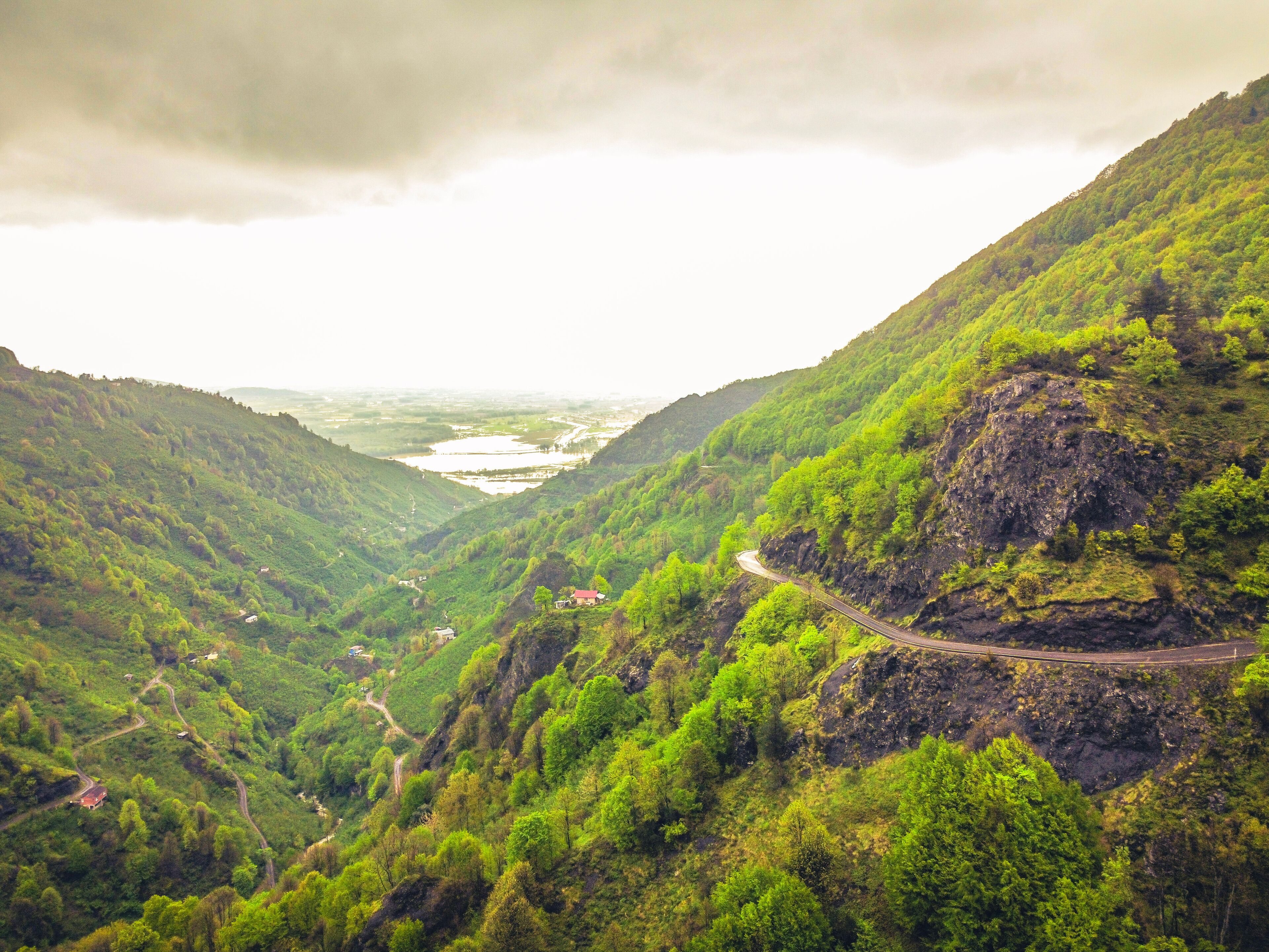 A winding mountain road through a lush valley with dense greenery and a river in the distance.