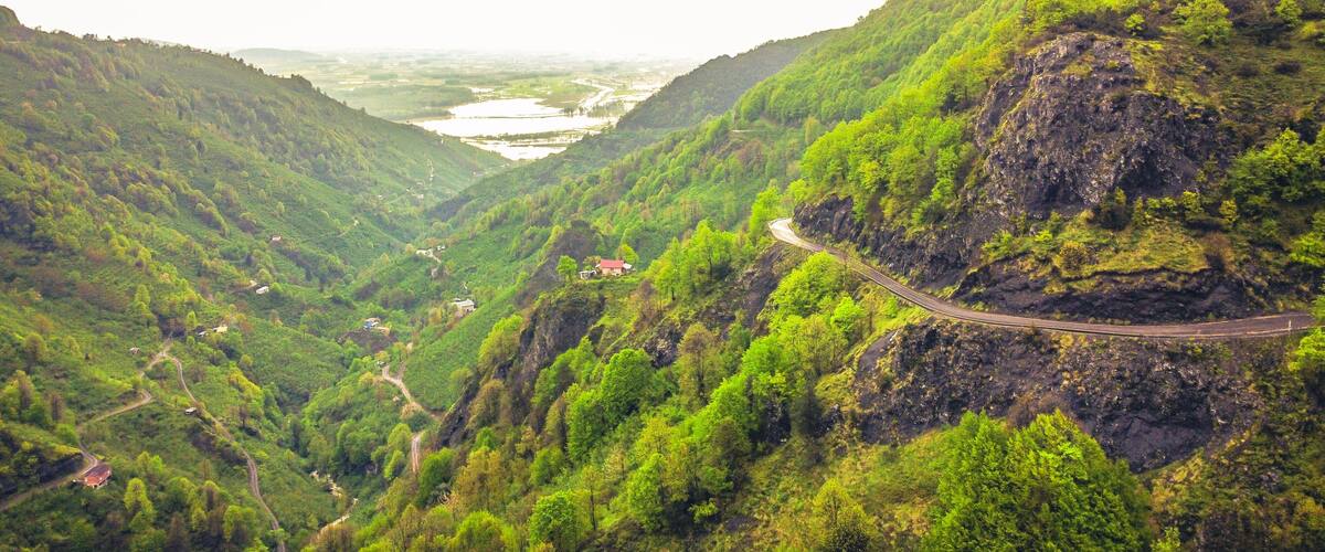A winding mountain road through a lush valley with dense greenery and a river in the distance.