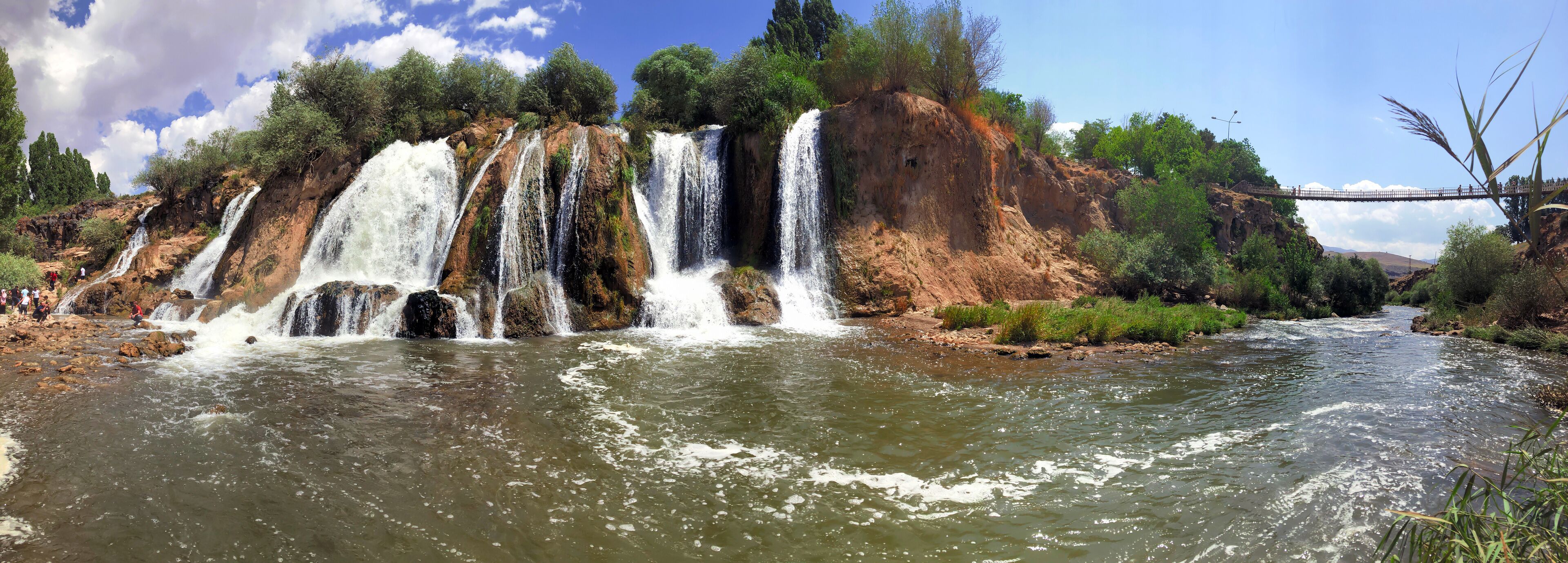 A multi-tiered waterfall cascading into a river surrounded by rocky terrain and greenery.