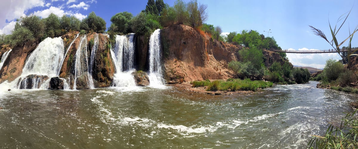 A multi-tiered waterfall cascading into a river surrounded by rocky terrain and greenery.