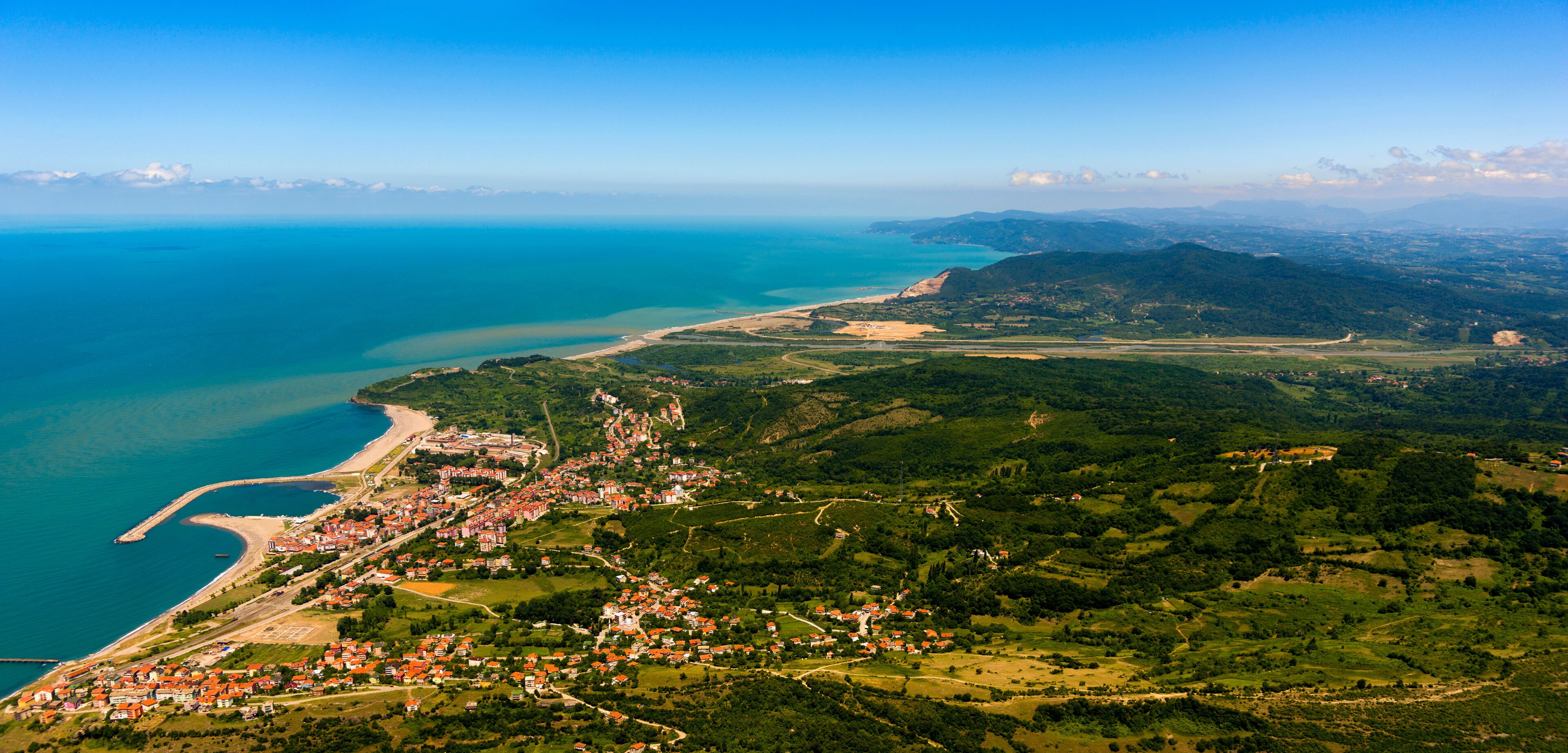 An aerial view of a coastal town with a beach, dense greenery, and a mountainous backdrop.