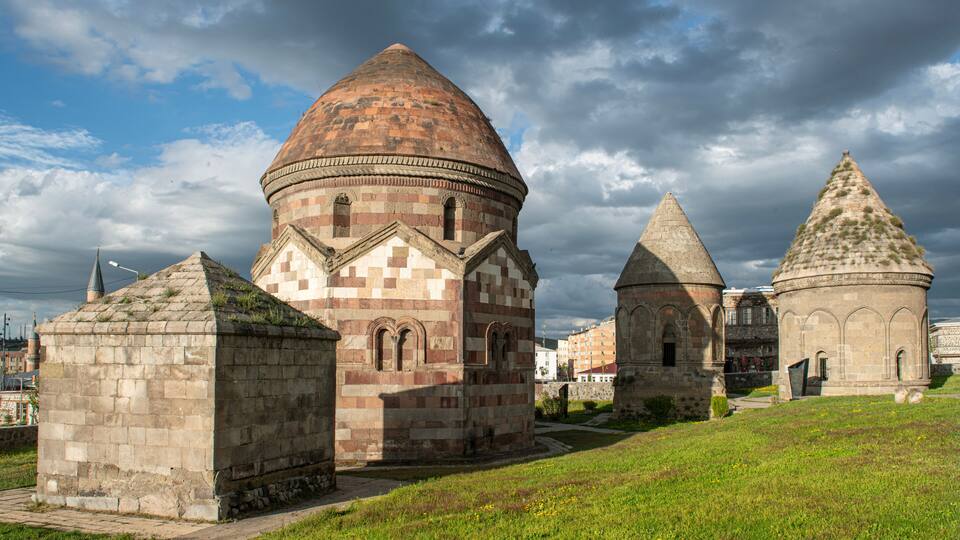 A historic building with a large dome and smaller structures, surrounded by green grass and under a cloudy sky.