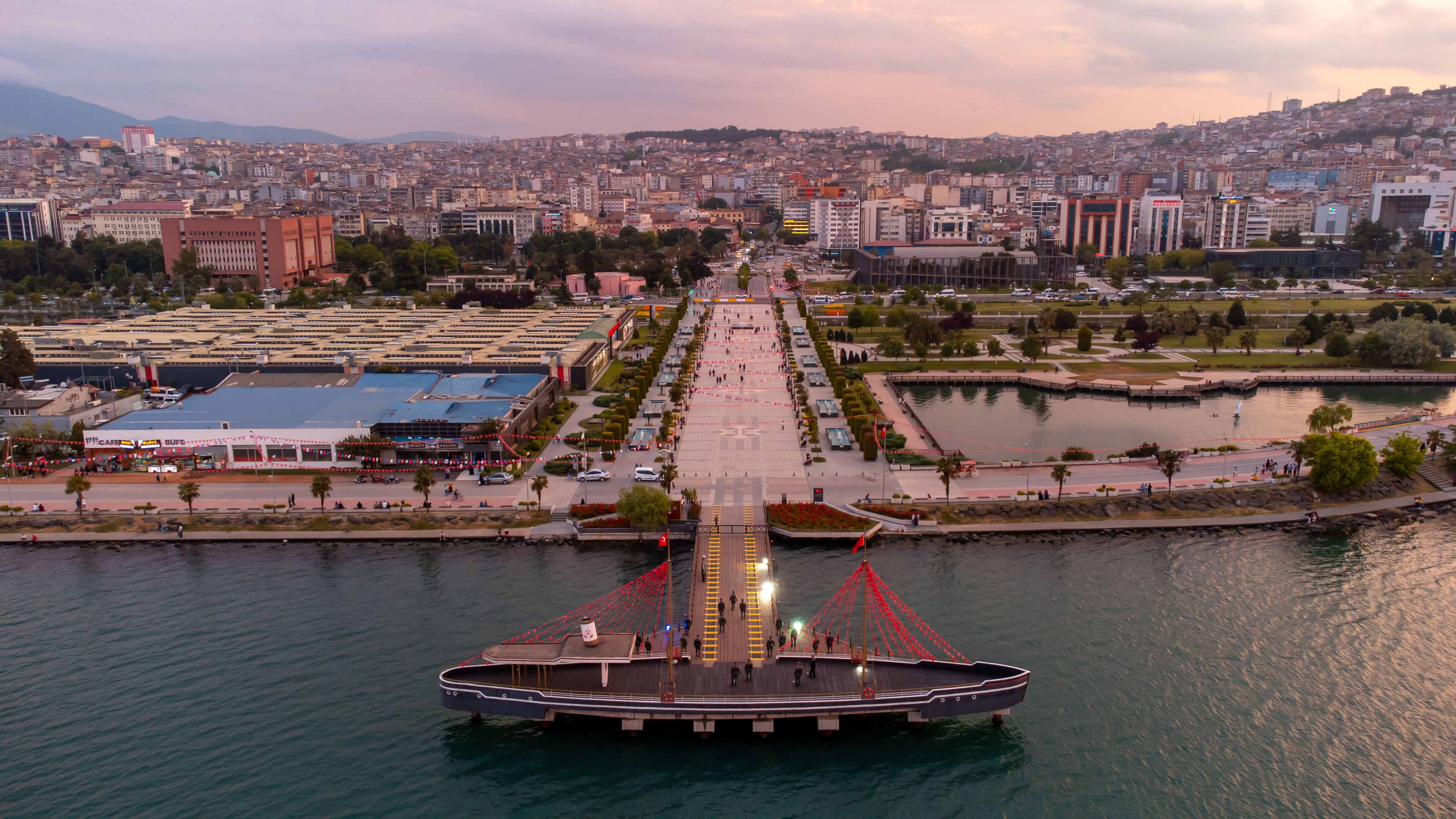 A waterfront promenade with a large boat dock, a cityscape in the background, and a river.