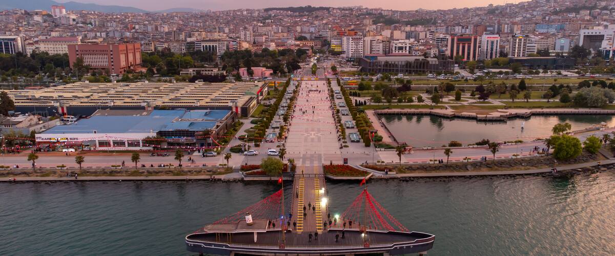 A waterfront promenade with a large boat dock, a cityscape in the background, and a river.