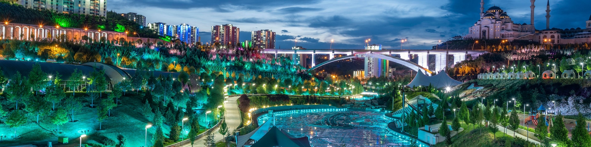 Vista urbana noturna com edifícios iluminados, um rio e uma mesquita.