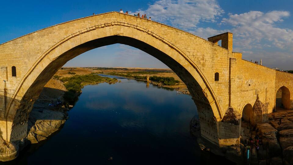 An ancient stone bridge arching over a calm river, surrounded by rocky terrain and a clear sky.