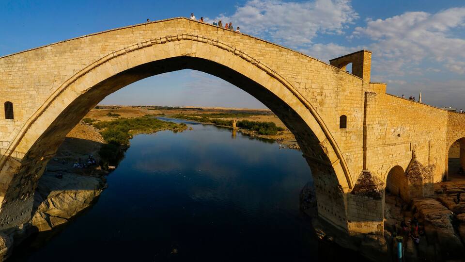 An ancient stone bridge over a calm river, surrounded by rocky terrain and historical ruins.