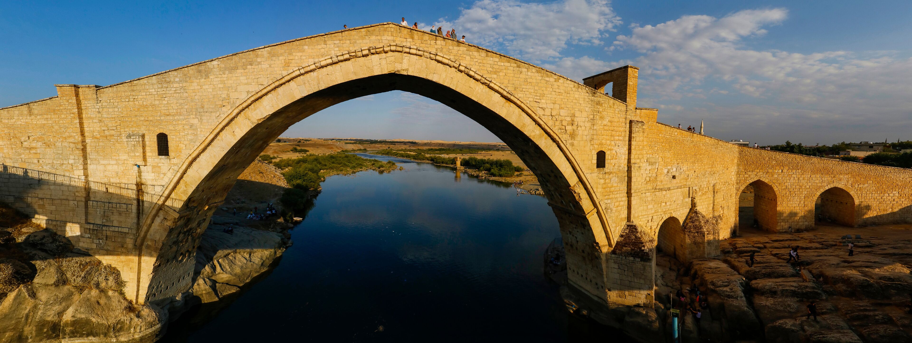 An ancient stone bridge over a calm river, surrounded by rocky terrain and historical ruins.