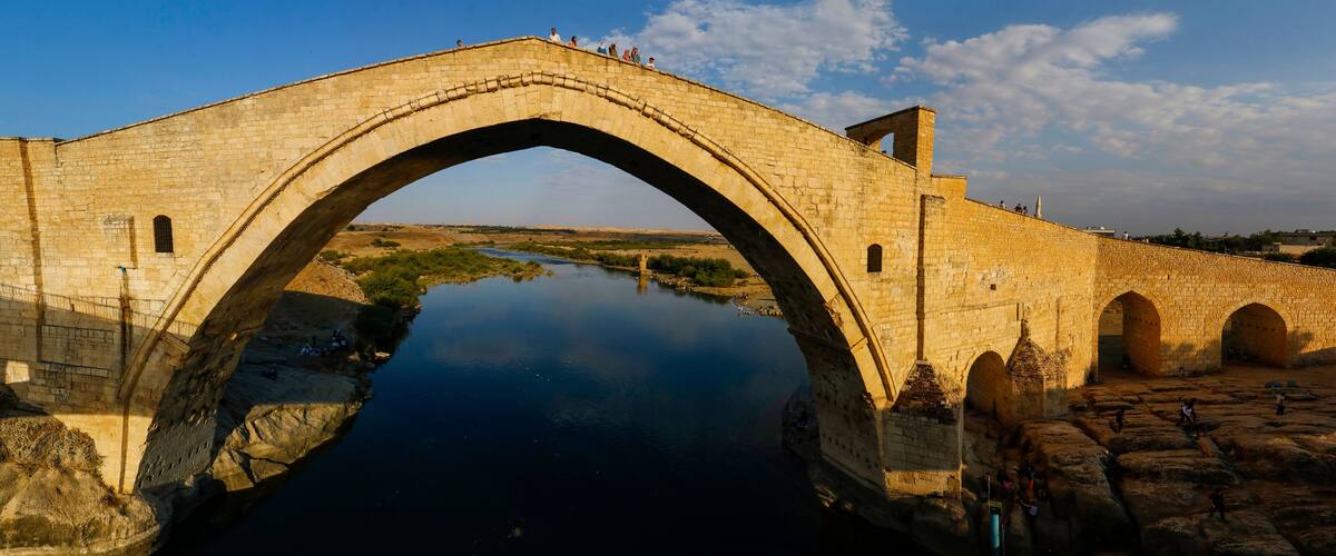 An ancient stone bridge over a calm river, surrounded by rocky terrain and historical ruins.