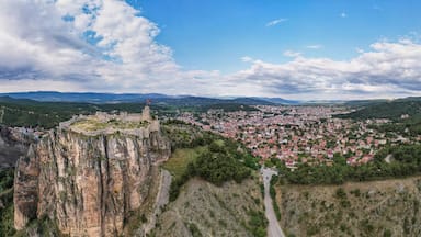 A town nestled in a valley with a prominent castle on a cliff, surrounded by mountains and a winding road.