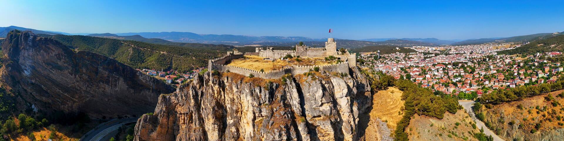 A panoramic view of Boyabat Castle perched on a dramatic rocky cliff in Sinop, Turkey, with the town in the background and forested hills under a vivid blue sky on a sunny day. Turkiye Turkey panorama