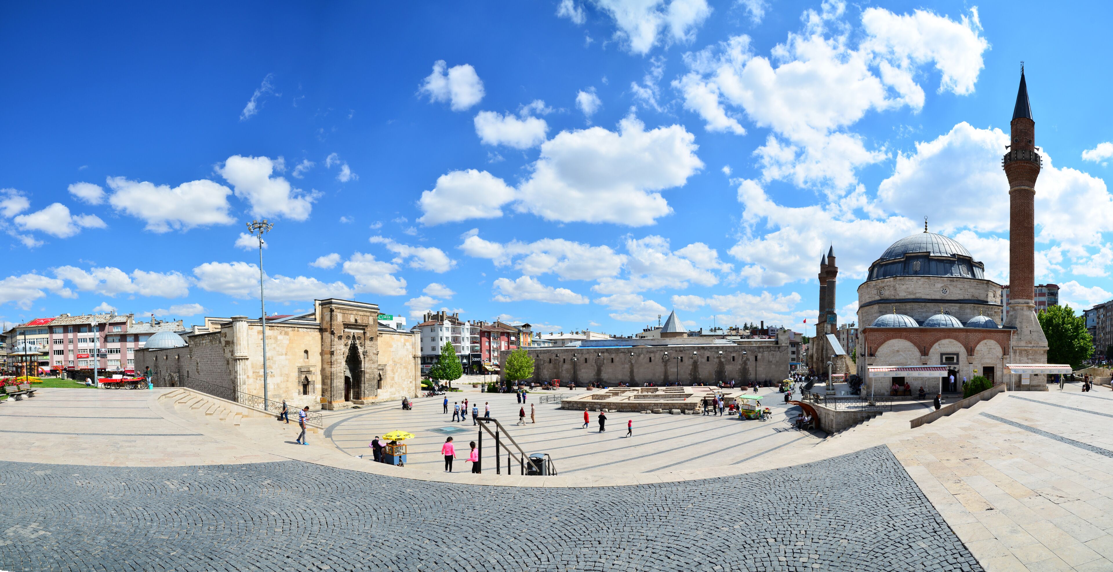 A historic square with a prominent mosque featuring a tall minaret and a dome, surrounded by stone buildings and cobblestone streets.