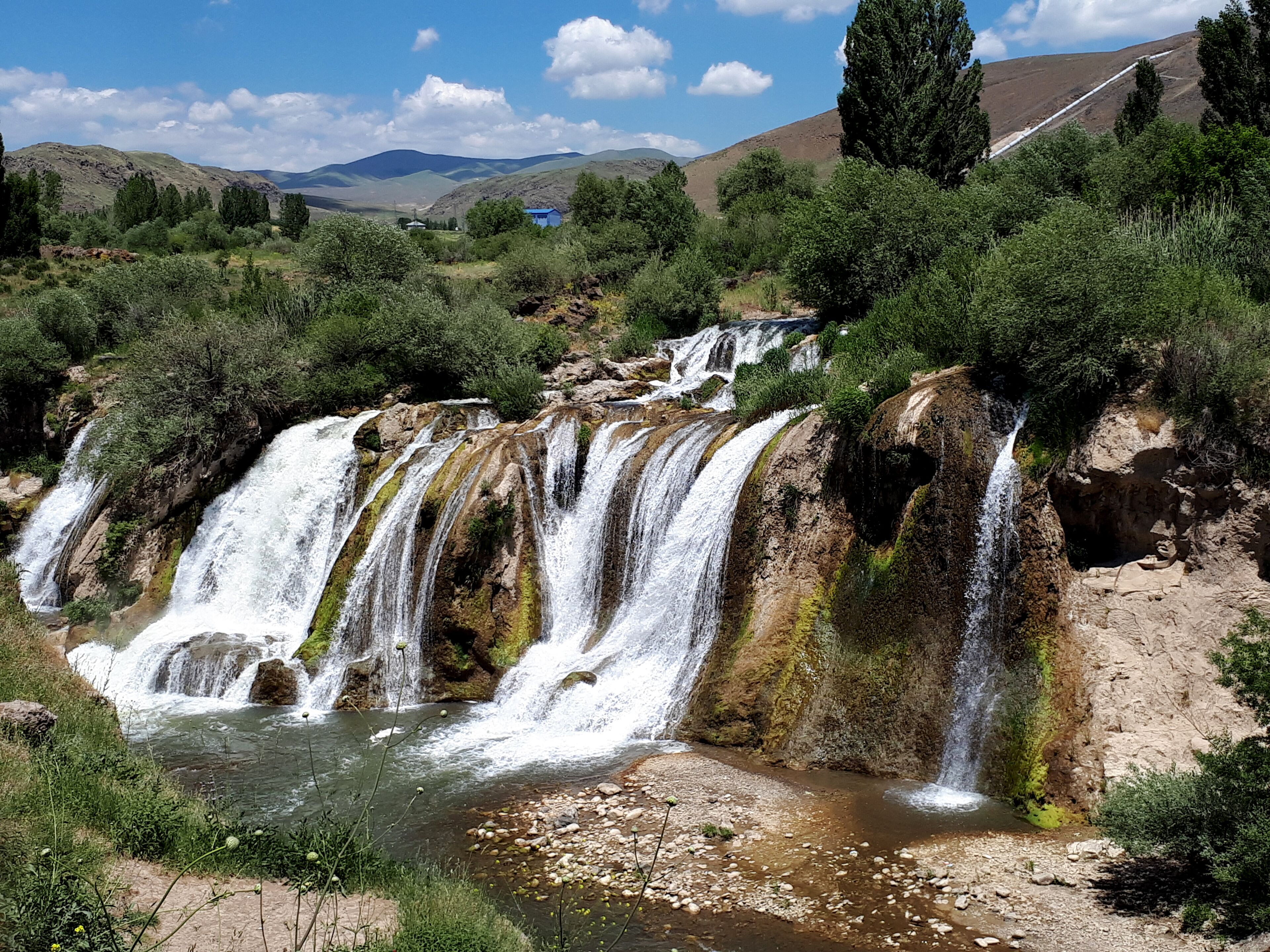 A multi-tiered waterfall in a lush valley with surrounding hills and sparse vegetation.