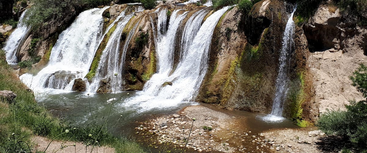 A multi-tiered waterfall in a lush valley with surrounding hills and sparse vegetation.