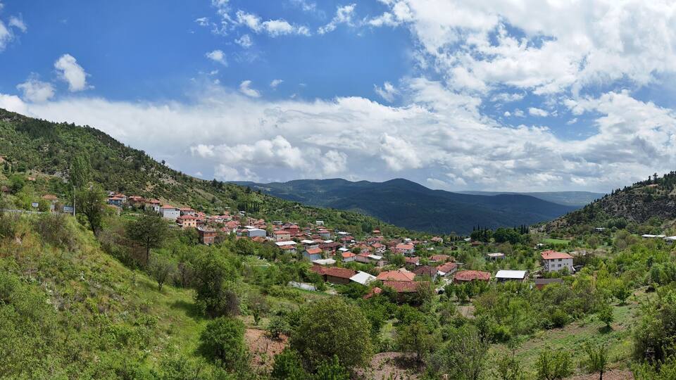 Panoramic view of Keles Kemaliye village in Turkey, showcasing green hills, traditional houses, and a dramatic cloudy sky, reflecting the peaceful rural life and natural beauty of the region.