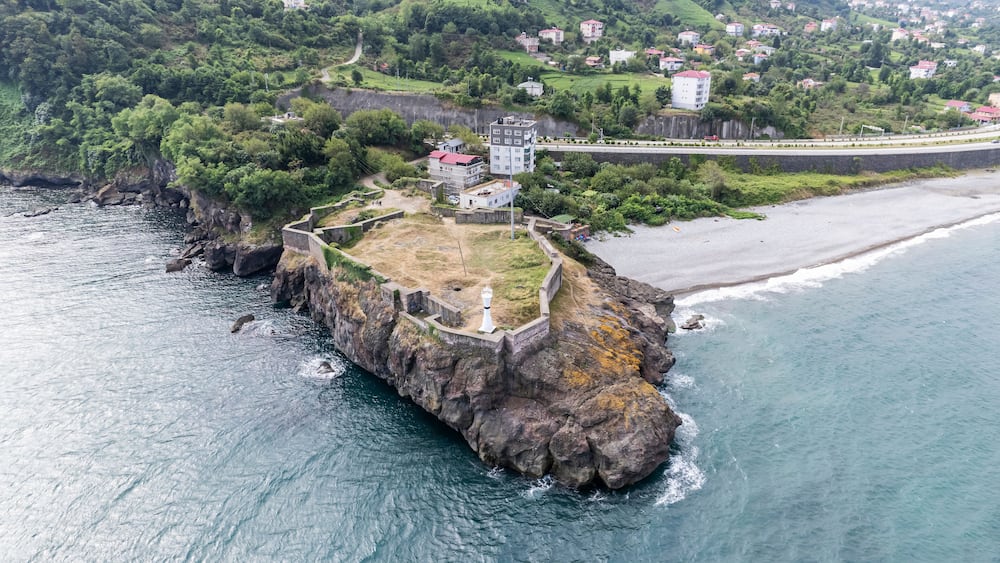 A coastal fortification with a lighthouse, surrounded by a rocky coastline and a residential area in the background.