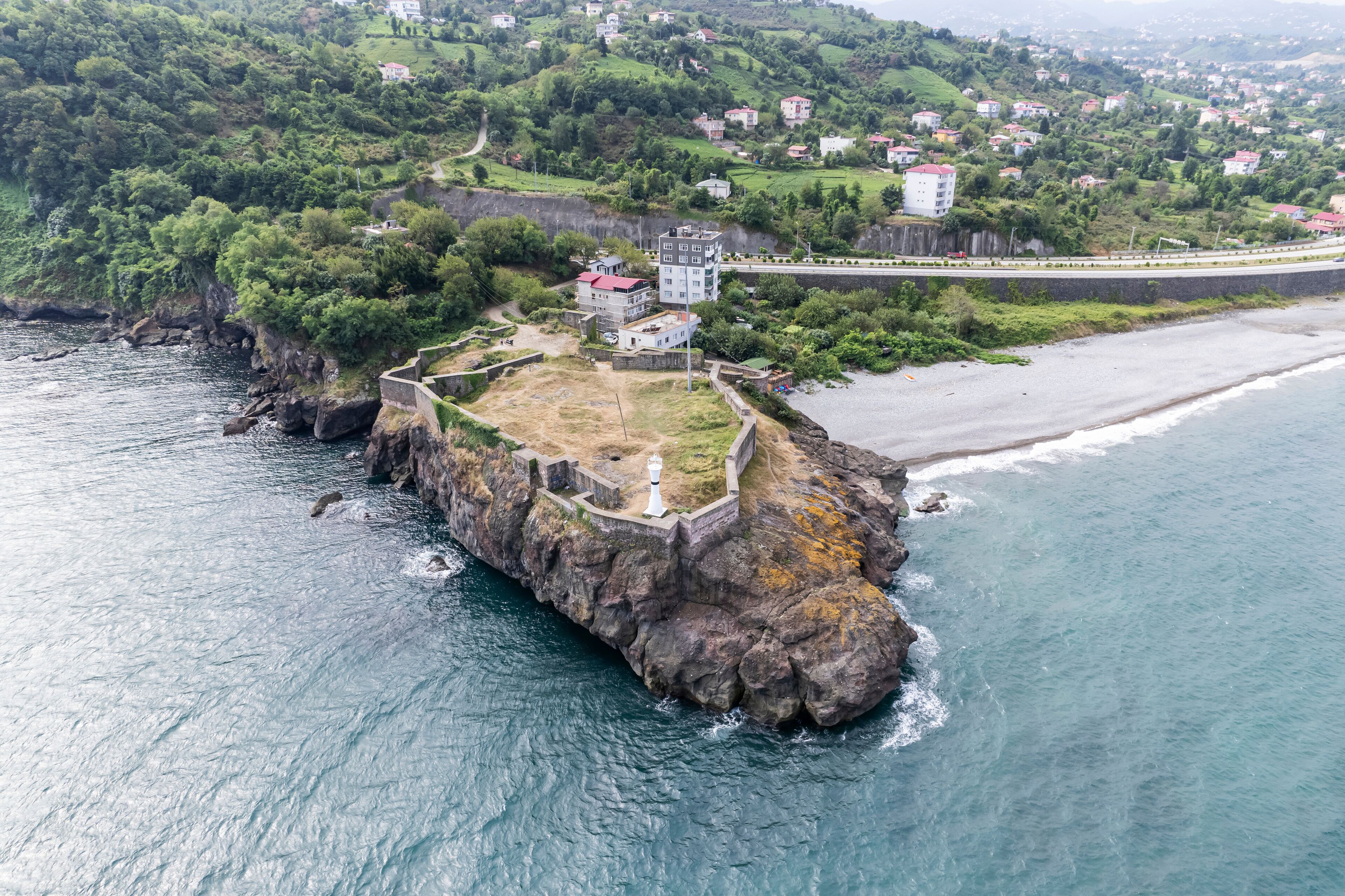 A coastal fortification with a lighthouse, surrounded by a rocky coastline and a residential area in the background.