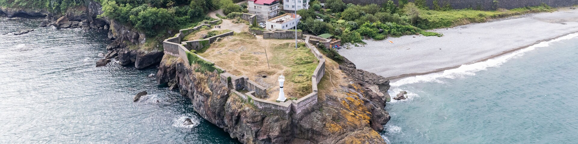 A coastal fortification with a lighthouse, surrounded by a rocky coastline and a residential area in the background.