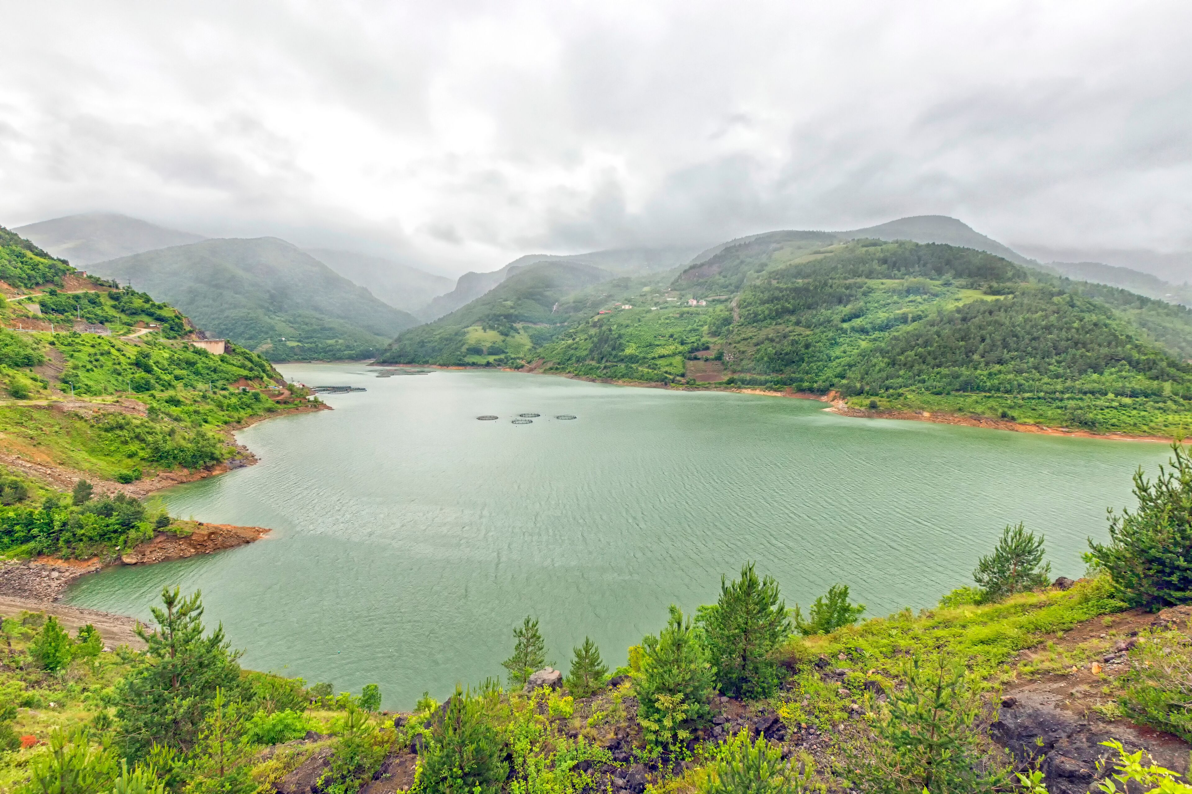 A calm lake surrounded by lush green hills and mountains under a cloudy sky.