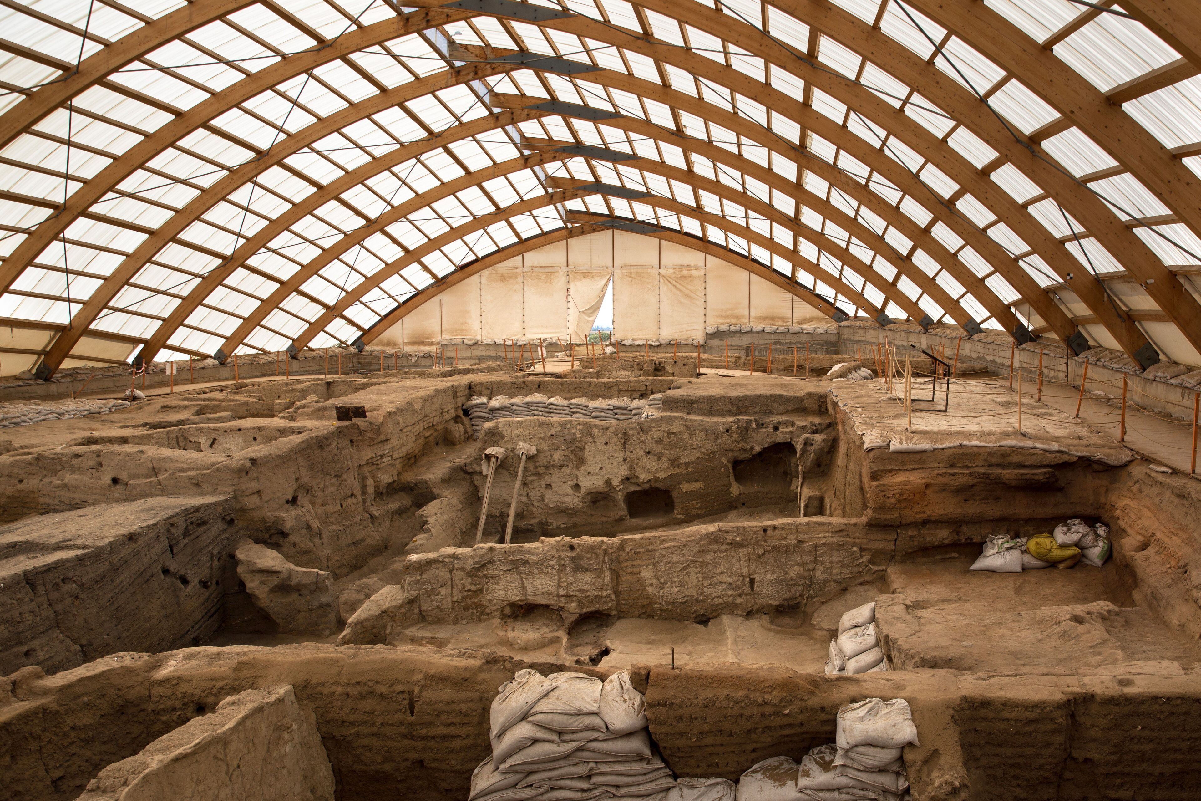 An archaeological site with a large, arched wooden roof and exposed stone structures.