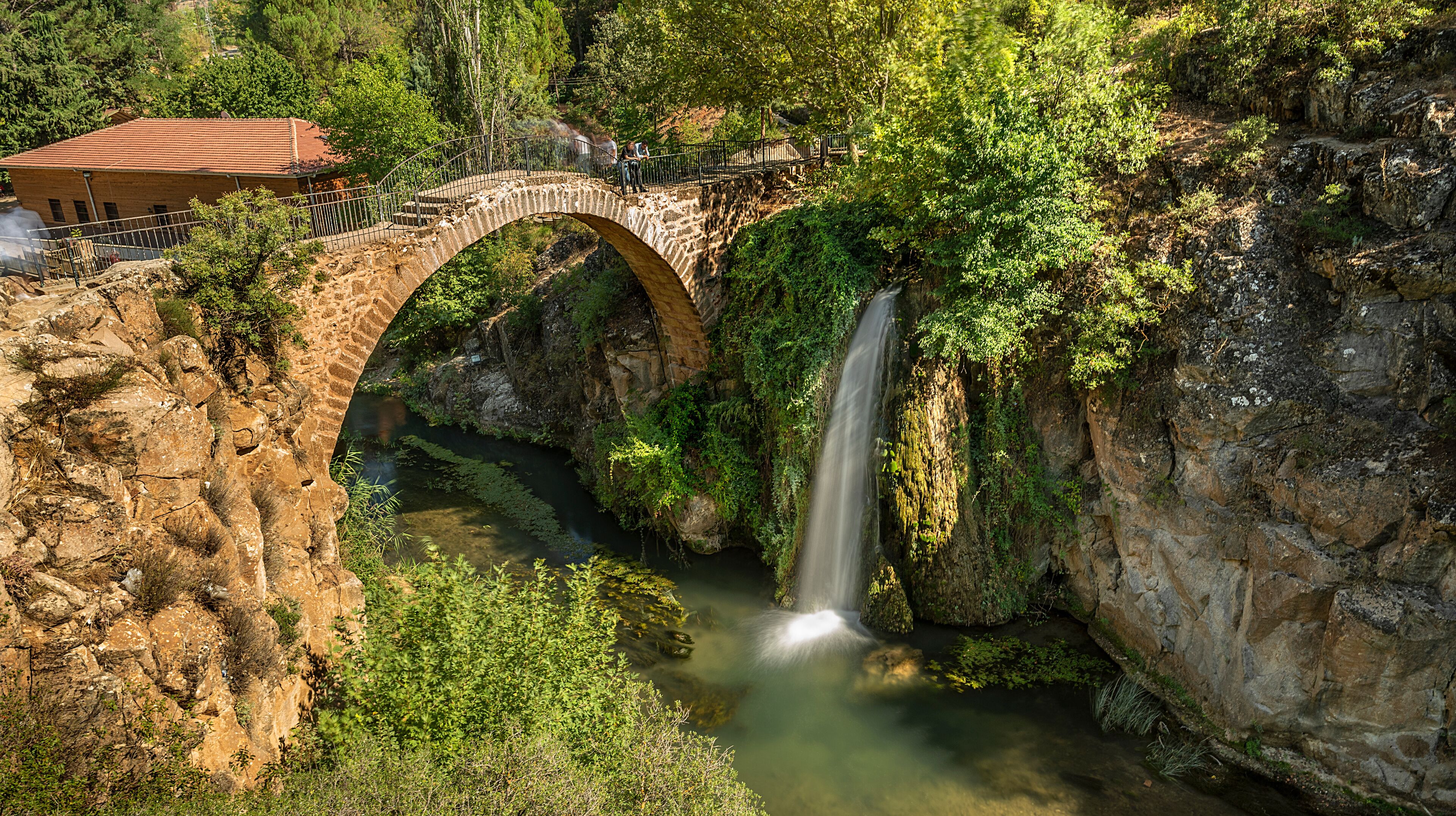 A stone bridge over a waterfall, surrounded by lush greenery and a building with a red roof.