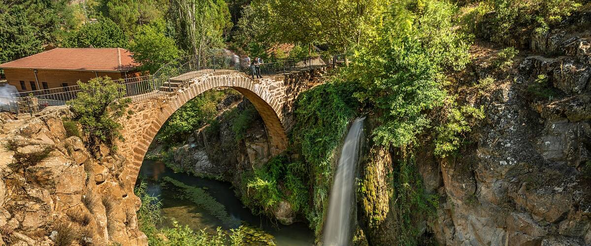 A stone bridge over a waterfall, surrounded by lush greenery and a building with a red roof.