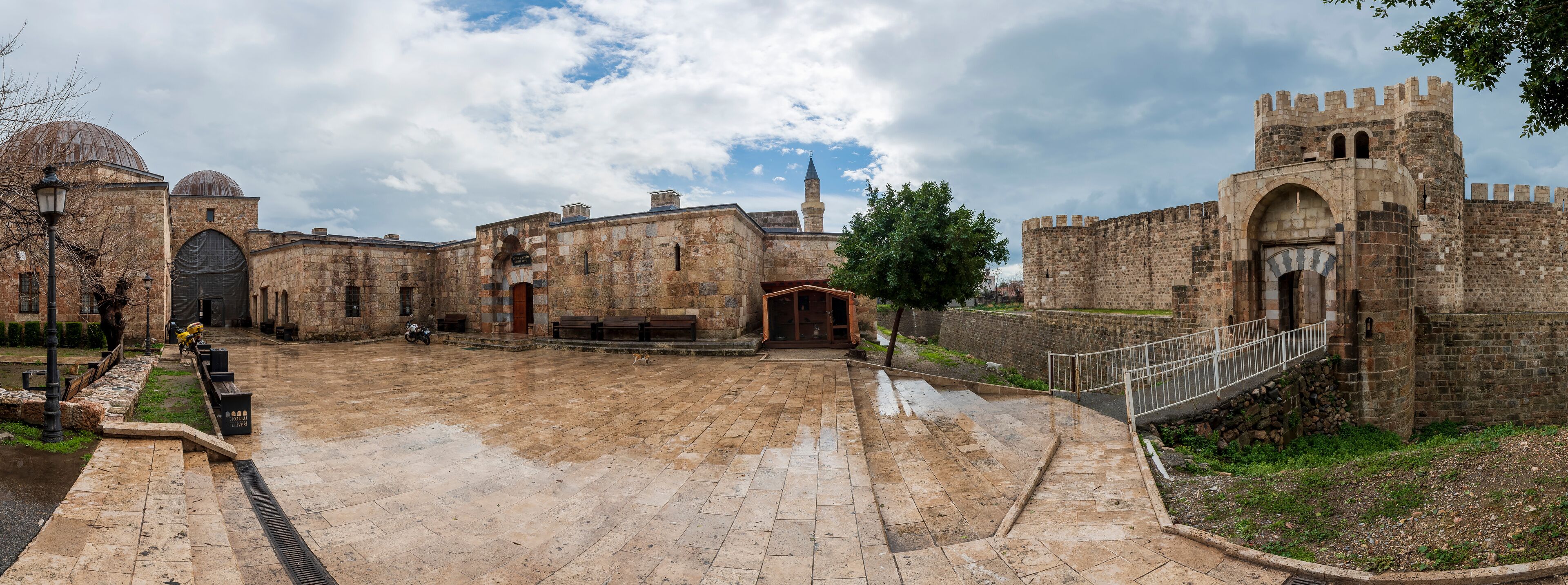 A historic courtyard with stone buildings, a mosque with a dome, and a fortified wall with a gate.