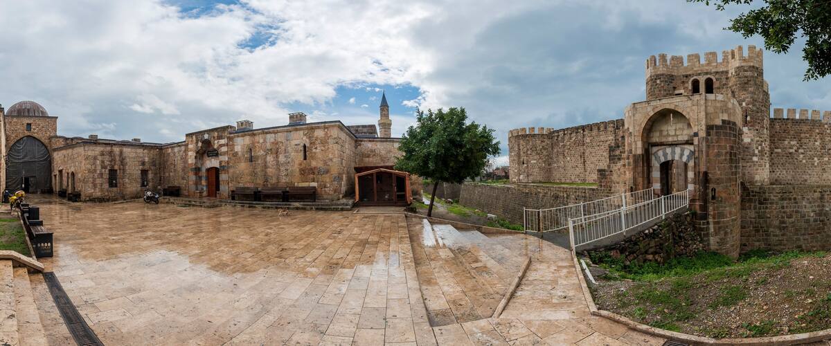 A historic courtyard with stone buildings, a mosque with a dome, and a fortified wall with a gate.