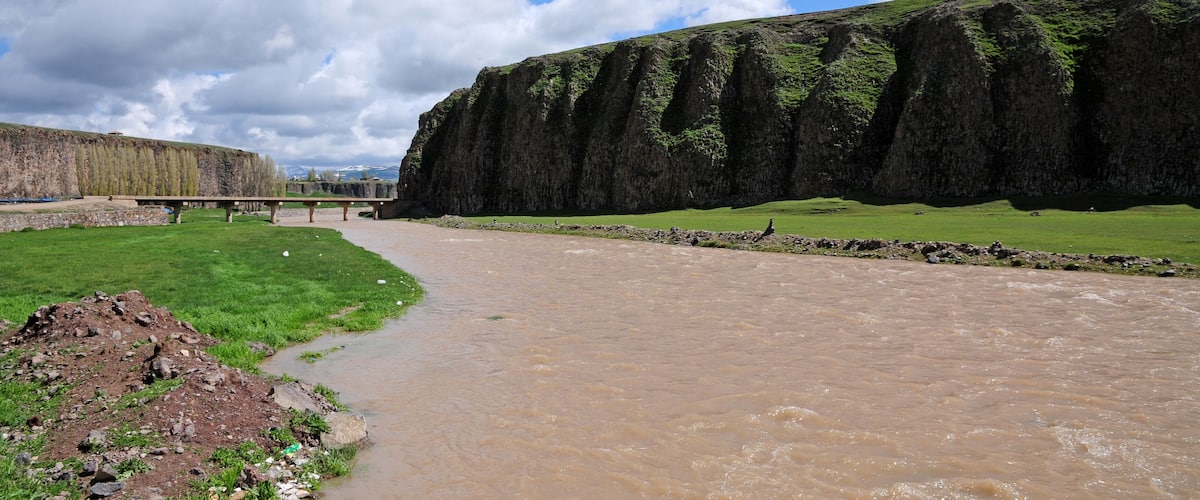 A river flowing through a green landscape with a rocky cliff and a bridge in the background.