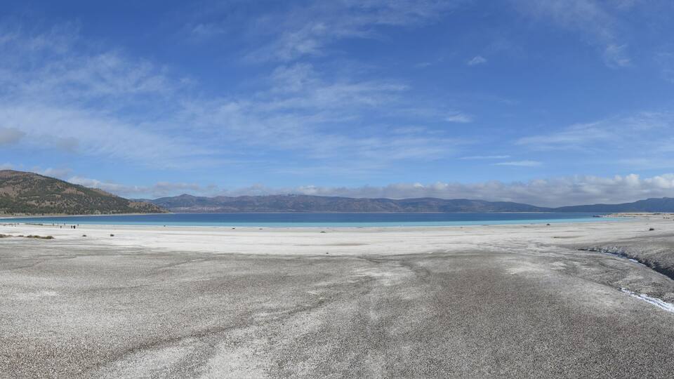 A coastal landscape with a pebble beach, a clear blue sea, and distant mountains under a partly cloudy sky.