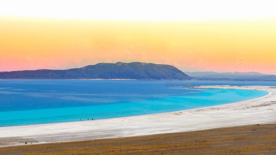 A coastal landscape with a white sandy beach, turquoise waters, and distant mountains under a sunset sky.