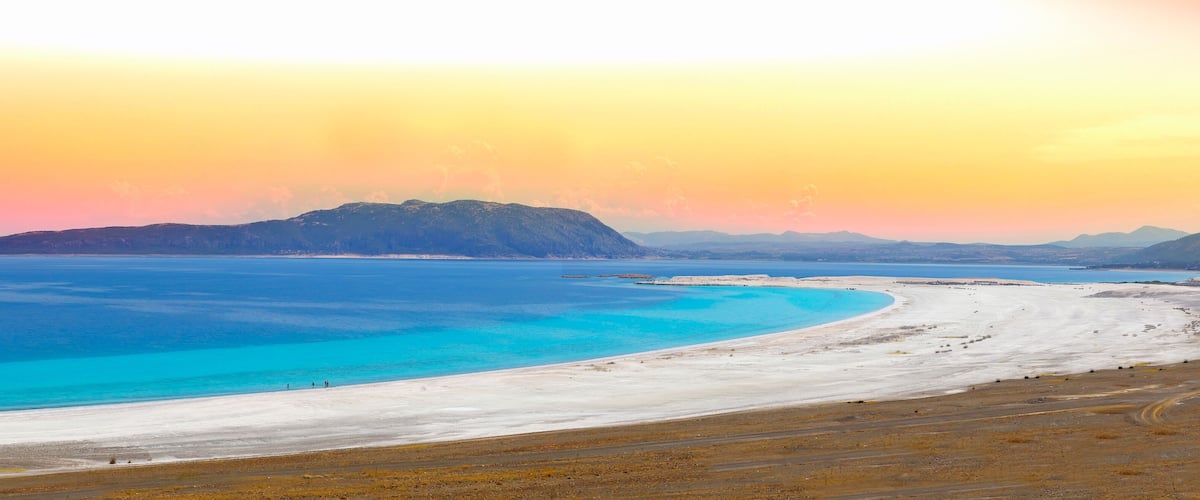 A coastal landscape with a white sandy beach, turquoise waters, and distant mountains under a sunset sky.