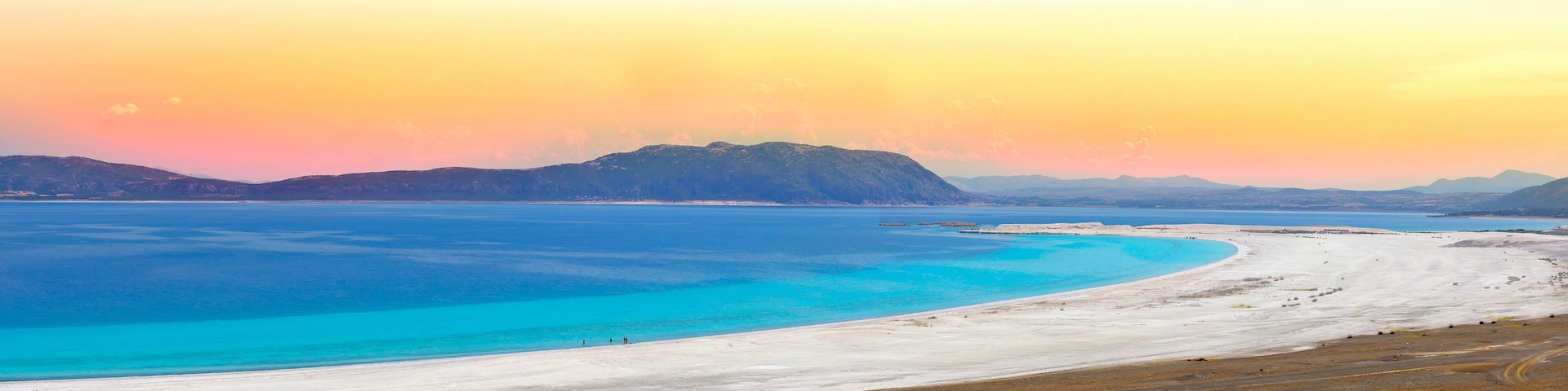 A coastal landscape with a white sandy beach, turquoise waters, and distant mountains under a sunset sky.