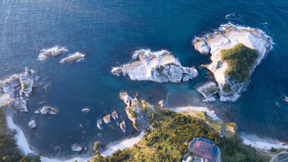 An aerial view of a coastal area with rocky formations, clear blue waters, and a small building on a grassy hill.